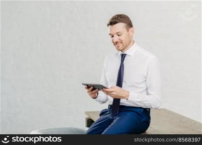 Positive male financier wears white shirt, tie, sits on table, installs new application on touch pad, connected to wireless internet. Successful businessman reads notification on modern device