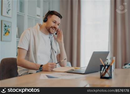 Positive male freelancer or student poses at home office wears headset takes part in education webinar makes notes looks gladfull at laptop display makes video call dressed casually. Web communication