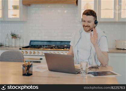 Positive male freelancer using laptop at home during online conference with colleagues while waving to them and looking at screen, sitting at his workplace at home office. Freelance concept. Happy young man having lesson during elearning on laptop at home