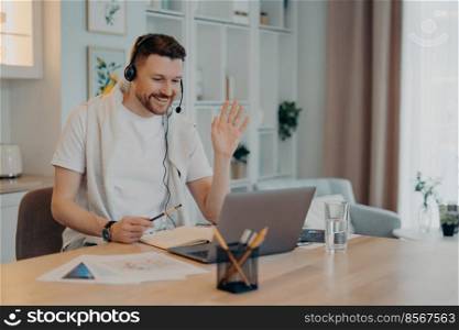 Positive male freelancer using laptop at home, sitting at wooden table and having online conference with colleagues while working remotely, waving to them. Freelance and remote job concept. Happy young guy in headphones working from home and using online communication