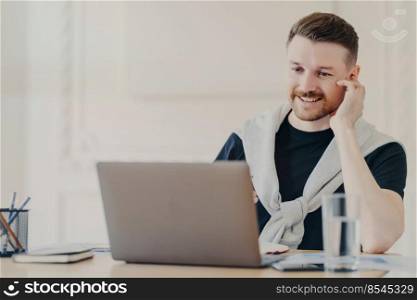 Positive male freelancer wearing casual clothes using earphones having online meeting, businessman smiling while sitting in front of laptop behind office desk and talking with colleagues online. Smiling male office worker having video call on laptop