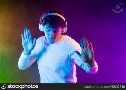 Positive man listening music with headphones, dancing on dark neon background. Stylish student guy enjoying life, active energy, inspired dance concept. High quality photo. Positive man listening music with headphones, dancing on dark neon background. Stylish student guy enjoying life, active energy, inspired dance concept