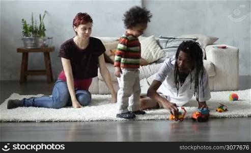 Positive mixed race family with adorable toddler son sitting on the floor in living room and playing together with car toys at home. Beautiful caucasian pregnant mother, african american father and their mixed race curly boy having fun and playing with toys.