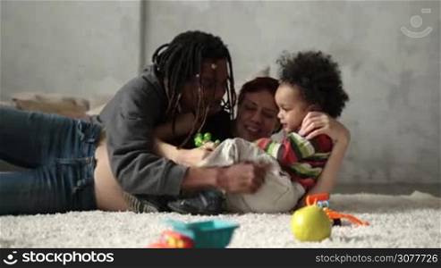 Positive multiethnic family and curly mixed race toddler playing on the floor in living room. Cheerful african american dad with dreadlocks, caucasian pregnant mother and little son lying on the carpet and having great time together. Slow motion.