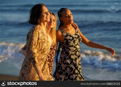 Positive multiracial female friends hugging and strolling together on seashore along waving sea on sunny summer day in coastal terrain. Young diverse girlfriends walking near sea