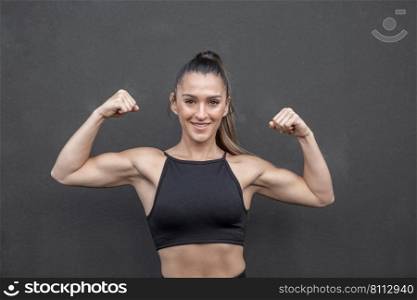 Positive muscular female athlete in black crop top looking at camera with smile and showing biceps while standing against black wall during workout in gym. Friendly sportswoman smiling and showing muscles