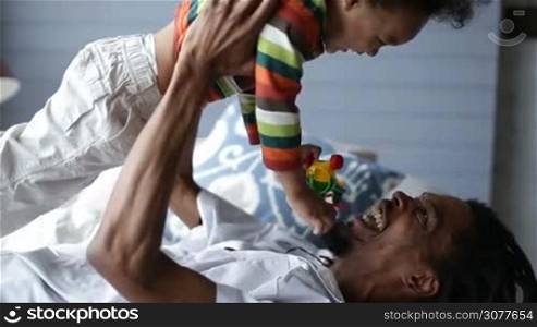 Positive playful african dad raising his cute toddler son up in the air while lying on his back on the bed at home. Handsome smiling father with dreadlocks playing with his mixed race boy in domestic interior. Side view closeup.