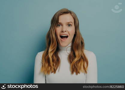 Positive pretty young woman with surprised shocked funny facial expression looking at camera with mouth wide open posing over blue studio wall background, portrait of amazed female. Amazed young woman with surprised funny facial expression looking at camera with mouth wide open