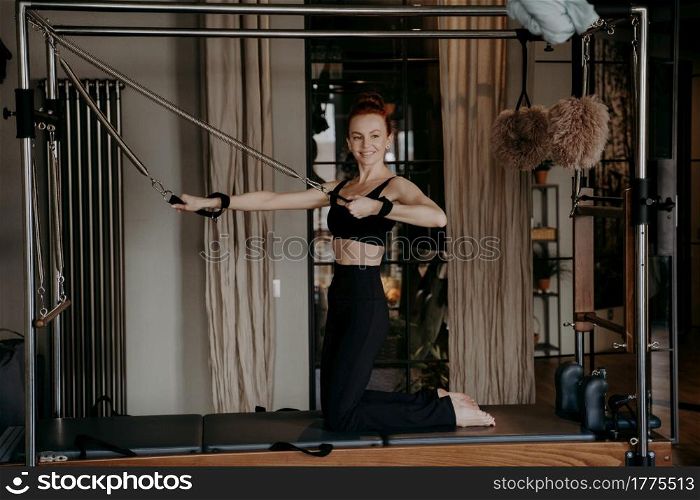 Positive red haired woman fit pilates instructor doing arm strength exercises on top of cadillac reformer happily smiling during training in gym or studio, using special fitness equipment. Happy healthy ginger woman doing arm strength exercises on reformer