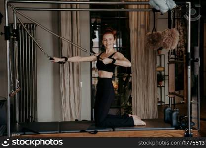 Positive red haired woman fit pilates instructor doing arm strength exercises on top of cadillac reformer happily smiling during training in gym or studio, using special fitness equipment. Happy healthy ginger woman doing arm strength exercises on reformer