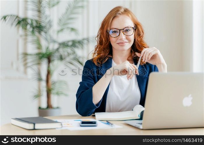 Positive red haired young female entrepreneur watches business webinar, writes email, makes notes in notepad, wears optical glasses, dressed in formal clothes, works online, involved in banking sphere