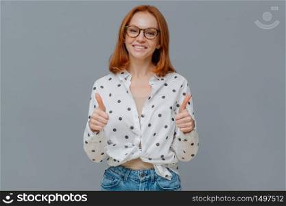 Positive redhaired woman with gentle smiles shows thumbs up, demonstrates like or approval gesture, says well done, likes someting, wears polka dot shirt and jeans, isolated on grey studio wall