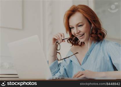 Positive redhead businesswoman participates in online conference, has training class with tutor, focused in display of laptop computer, holds spectacles, enjoys watching educational webinar.