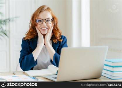 Positive redhead executive manager searches information on internet website, touches both cheeks, writes notes in notepad, uses modern laptop computer, dressed in formal clothes, checks organiser.