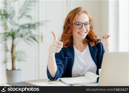 Positive redhead woman keeps thumb raised, demonstrates like gesture, satisfied with good work of colleague, updates software on modern gadget, searches data on website, poses in coworking space