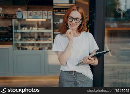 Positive redhead woman uses earphones with built in microphone tablet to communicate with friends and colleagues has conversation via internet wears spectacles white t shirt grey formal trousers. Positive redhead woman uses earphones with built in microphone tablet to communicate