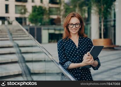 Positive redhead young woman in stylish outfit carries notepad poses outdoor against blurred background looks directly at camera. Pretty ginger female model wears polka dot dress walks at street. Positive redhead young woman in stylish outfit carries notepad poses outdoor
