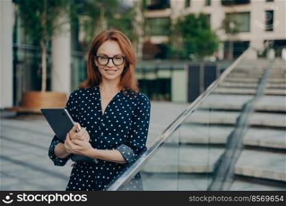 Positive redhead young woman in stylish outfit carries notepad poses outdoor against blurred background looks directly at camera. Pretty ginger female model wears polka dot dress walks at street. Positive redhead young woman in stylish outfit carries notepad poses outdoor
