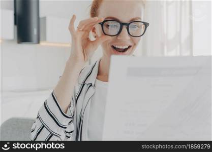 Positive redhead young woman smiles broadly wears transparent glasses focused at papers checks financial report glad to finish work in time works from home. People paperwork and job concept.. Positive redhead young woman smiles broadly wears transparent glasses focused at papers
