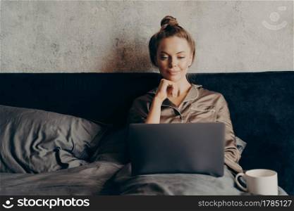 Positive relaxed young woman surfing internet for favorite film to watch on laptop while spending lazy day off at home, lying in bed at home and looking at computer monitor, dressed in pajama. Positive relaxed young woman in pajama lying in bed and using laptop