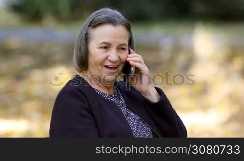 Positive senior woman talking on smartphone outdoors in park.