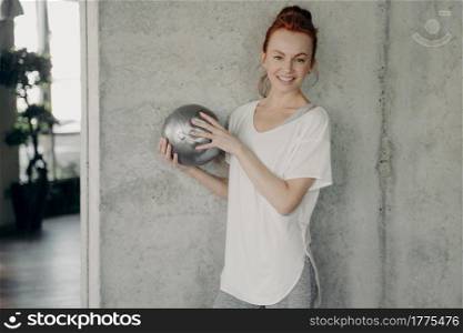 Positive slim young female with foxy hair stands by wall in white sport t shirt and gray leggings, holding small silver fitball by both hands and smiling on camera while exercising in fitness studio. Pretty slim woman with foxy hair stands by wall holding small silver fitball by both hands