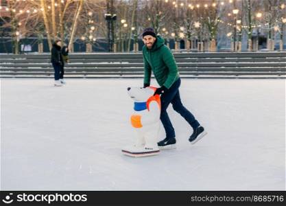 Positive smiling attractive man in warm winter clothes and skates, practices go skating on ice, uses skate rink, looks delightfully into camera, spends Sunday outdoor during winter period