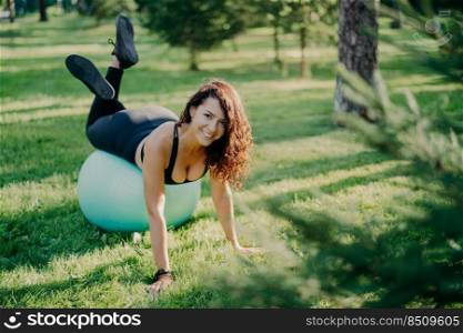 Positive smiling brunette woman exercises with fitness ball on green grass wants to have sporty body has curly hair dressed in sportswear has yoga training outdoor in park during warm sunny day