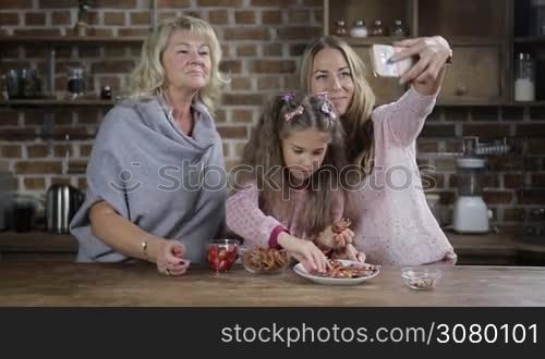 Positive smiling grandmother, mother and cute granddaughter taking selfie with smartphone while sitting at the kitchen table. Cheerful multi generation family making self portrait photo on mobile phone and having fun at home. Slow motion.