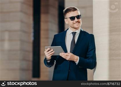 Positive smiling male manager with stubble, wears sunglasses and black suit, holds modern touch pad, checks email in internet, recieves positive news, looks into distance. People, business and style