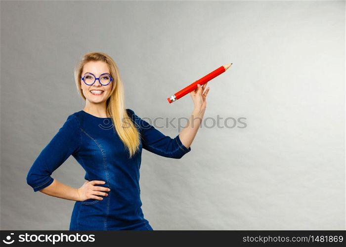 Positive smiling woman blonde student girl or female teacher holding big red pencil drawing. Studio shot on grey.. Smiling woman holds big pencil in hand