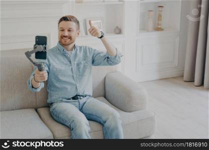 Positive smiling young man dressed in casual clothes showing thumbs up gesture in video chat, holding phone using gimbal, sitting alone on beige couch in light themed living room. Young man showing thumbs up gesture on camera on mobile phone
