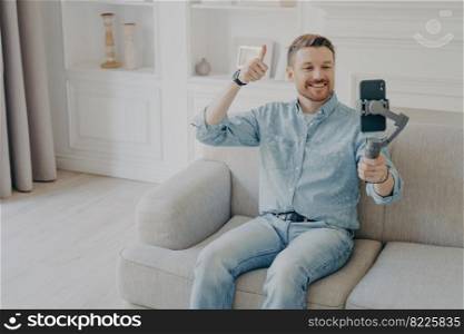 Positive smiling young man dressed in casual clothes showing thumbs up gesture in video chat, holding phone using gimbal, sitting alone on beige couch in light themed living room. Young man showing thumbs up gesture on camera on mobile phone