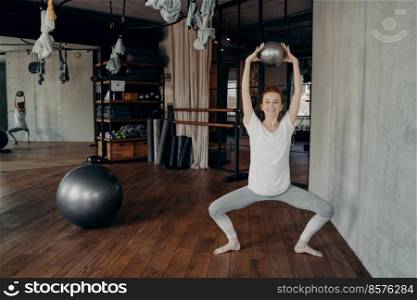 Positive sportive woman in good physical shape doing barre workout in fitness studio, sitting in plie and holding small fitball with both hands above her head in position with knees widely apart. Happy sportive female in active wear holding mini fitball above head in half sitting position