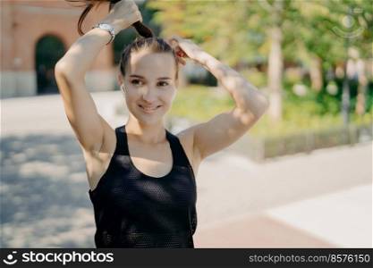 Positive sportswoman combes pony tail being in good physical shape listens music in earphones wears casual t shirt looks somewhere poses outdoor enjoys good weather sunny day. Sport concept.