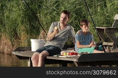 Positive stylish dad and teenage boy having picnic on wooden pontoon in summer sunlight while fishing together by the lake. Jouful father and son enjoying meal and communicating with each other. Slow motion.