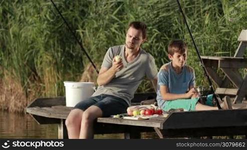 Positive stylish dad and teenage boy having picnic on wooden pontoon in summer sunlight while fishing together by the lake. Jouful father and son enjoying meal and communicating with each other. Slow motion.