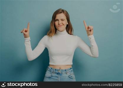 Positive stylish millennial girl in casual outfit pointing indicating with forefingers upwards at copy space and winking, promoting advertising product, standing isolated over blue studio background. Millennial girl pointing indicating with forefingers upwards and winking at camera
