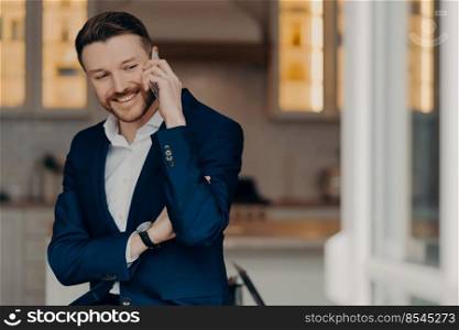 Positive successful businessman in suit standing in living room at home and enjoying talk on mobile phone with partner, holding smartphone and talking about job. Business communication concept. Happy executive director in stylish suit and with phone indoors