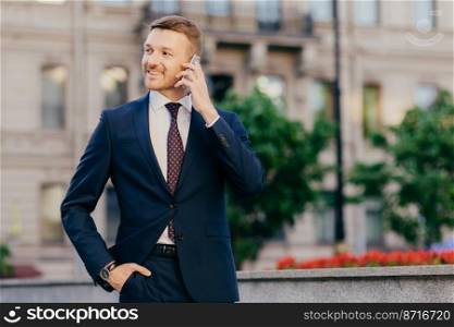 Positive successful businessman shares news with friends, glad to accomplish new innovative project, stands outdoor, keeps hand in pocket, dressed formally, notices something into distance aside