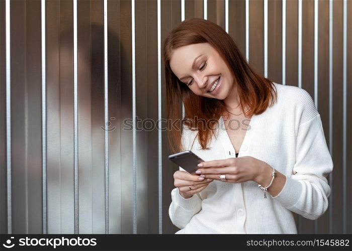 Positive successful European female economist makes money payment on website, poses with cell phone, dressed in white outfit, smiles happily, sends text message, enjoys online communication.