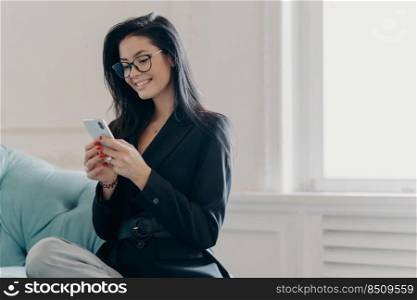 Positive successful female freelancer poses on comfortable sofa in living room, uses mobile phone, checks income message, types message, dressed in formal clothes, uses free internet connection