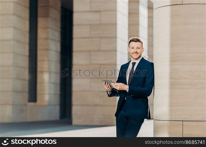 Positive successful male entrepreneur in luxury black suit, holds modern tablet, looks happily somewhere, stands in modern office building, searches internet or social networks during work break