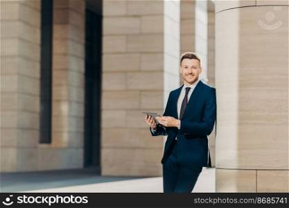 Positive successful male entrepreneur in luxury black suit, holds modern tablet, looks happily somewhere, stands in modern office building, searches internet or social networks during work break