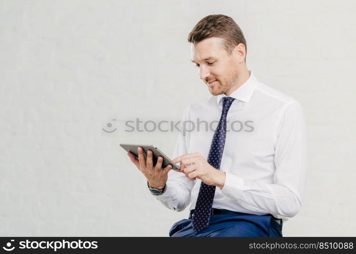 Positive successful male executive manager holds modern touch pad, gets notification about net feeds in social networks, satisfied with high speed internet connection, dressed in formal shirt with tie