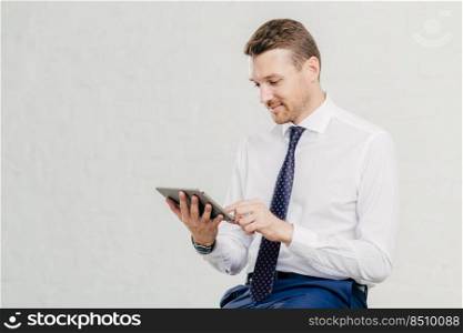 Positive successful male executive manager holds modern touch pad, gets notification about net feeds in social networks, satisfied with high speed internet connection, dressed in formal shirt with tie