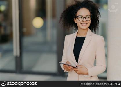Positive successful woman entrepreneur with Afro hair holds digital tablet, stands outdoor near office building, wears formal clothes, looks away, waits for colleague to have dinner during break