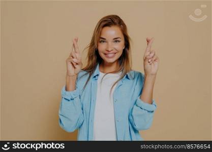 Positive superstitious young lady with beautiful smile holding her fingers crossed, making believer gesture, hoping for good luck and fulfillment of desire on beige background. Positive lady with fingers crossed on beige background