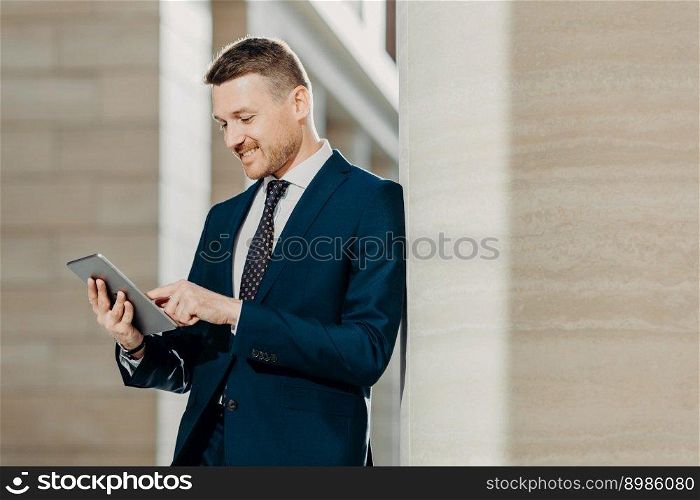 Positive unshaven businessman reads news in internet on web page, uses touch pad, dressed in elegant suit, has cheerful expression. Male executive enjoys communication with business partner online