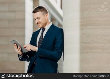 Positive unshaven businessman reads news in internet on web page, uses touch pad, dressed in elegant suit, has cheerful expression. Male executive enjoys communication with business partner online